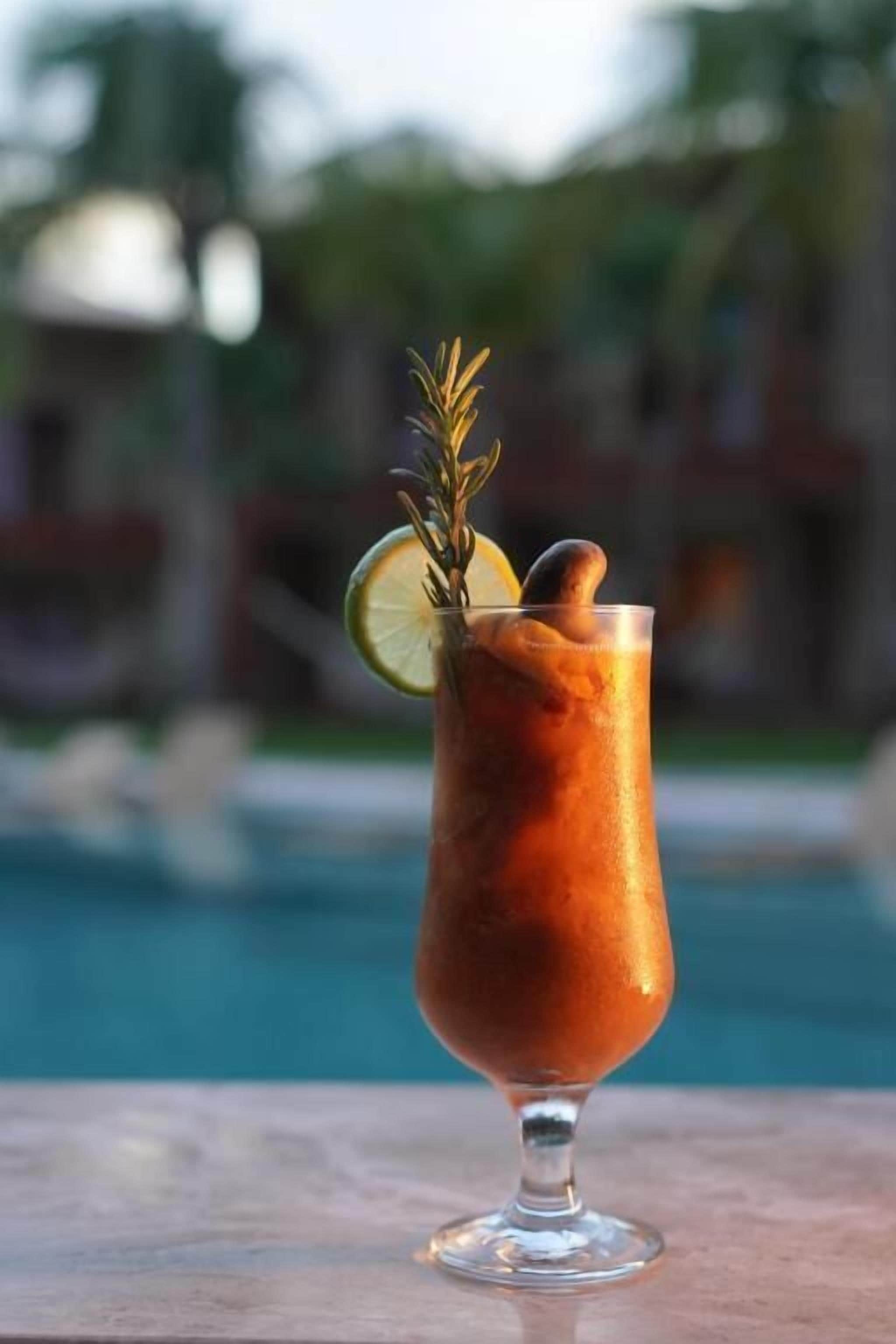 A cocktail in a stemmed glass with a lemon slice and a sprig of rosemary, placed on a table by a pool.