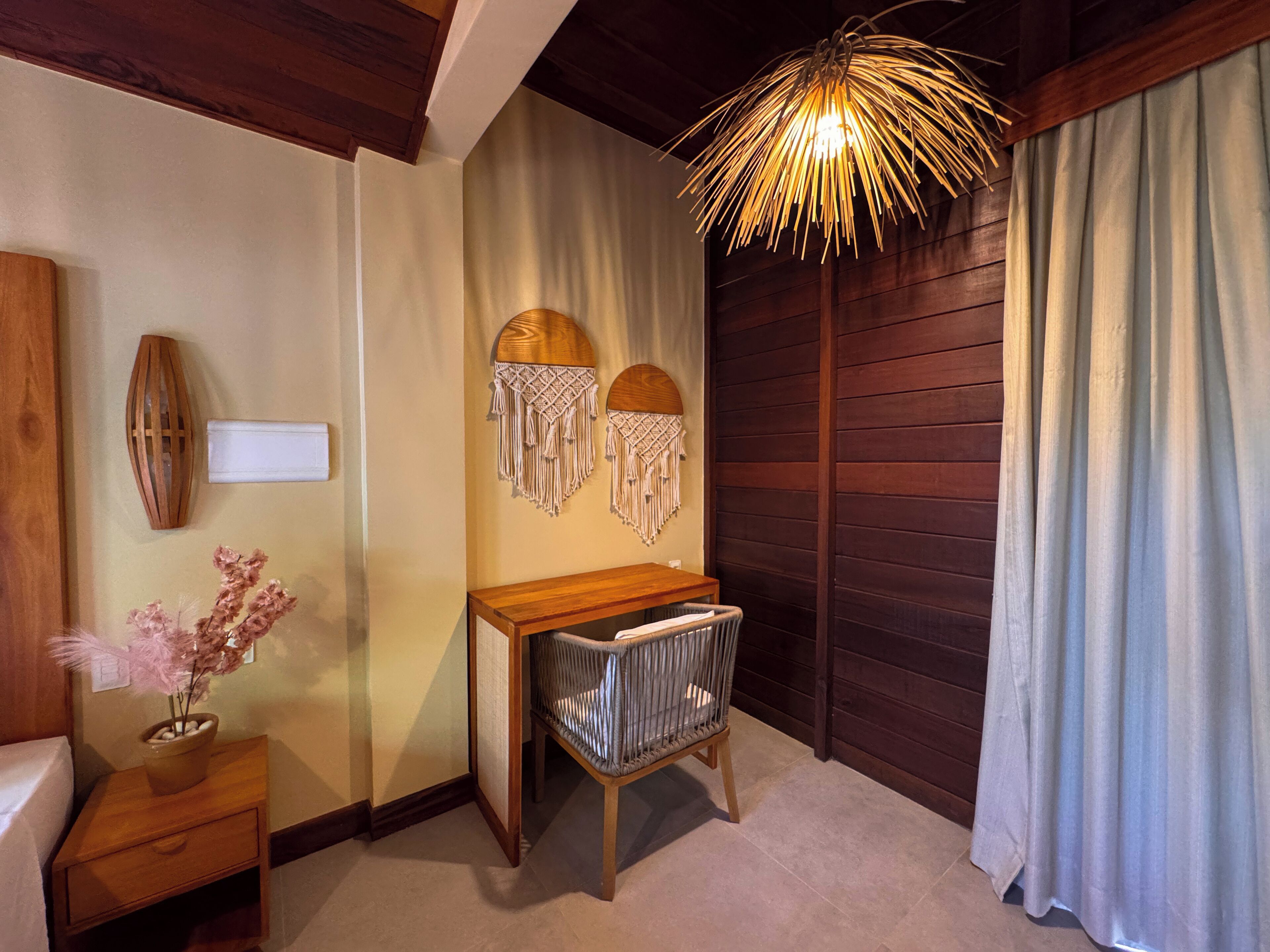 A room with a wooden desk, a woven chair, a macrame wall hanging, a vase with dried flowers, and a unique ceiling light fixture.