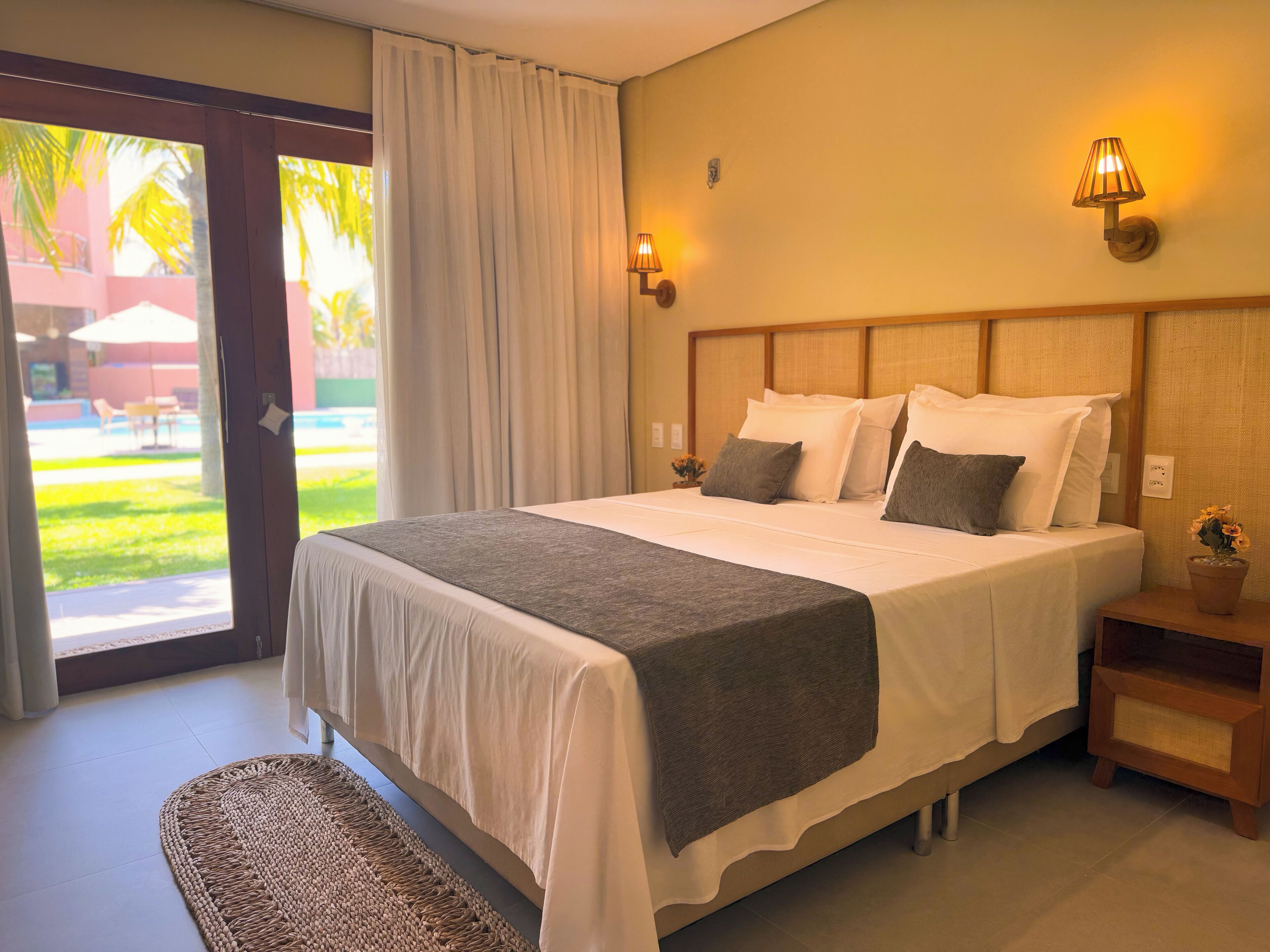 Une chambre d’hôtel avec un grand lit, une petite table de chevet avec une lampe, et une vue sur une piscine et des palmiers depuis la porte.