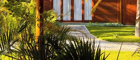 A wooden house with a thatched roof, surrounded by lush greenery and palm trees.