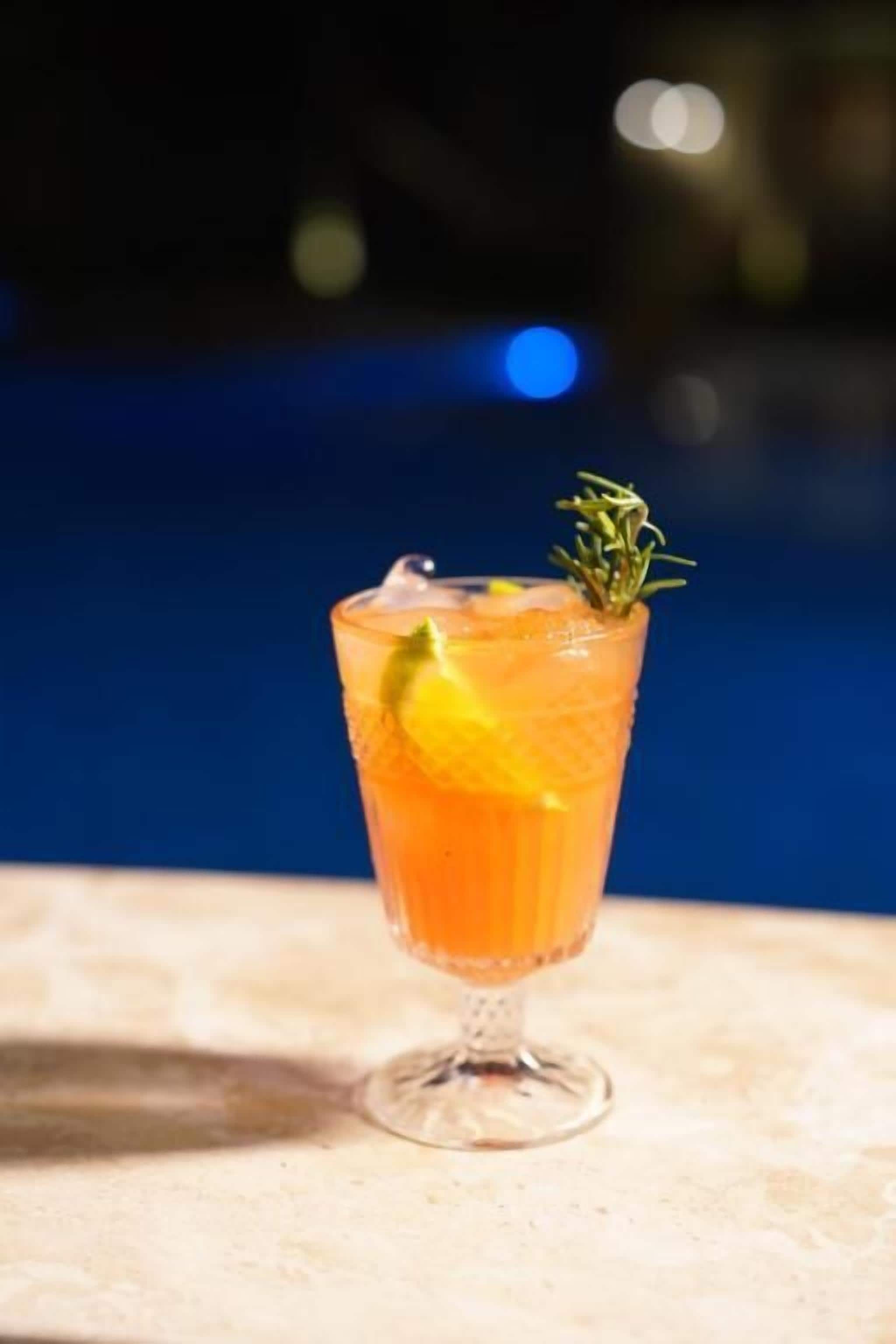 Un verre à cocktail, orné d’une tranche de citron et d’une branche de romarin, est posé sur une table au bord d’une piscine.