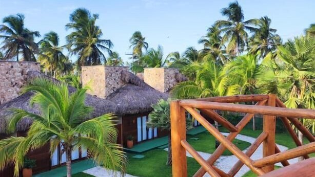 Un pontile di legno che si affaccia su un resort tropicale con edifici a tetto di paglia e palme.