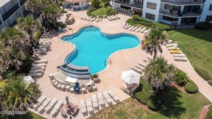 A swimming pool with lounge chairs and a small building in the center, surrounded by palm trees and a multi-story building with balconies.