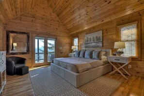 A log cabin bedroom with a large bed, a chair, a desk, and a window with blinds.