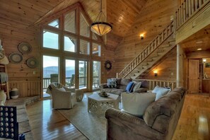 A spacious wooden cabin interior with a large window, a staircase, and a cozy seating area. - High Hopes | Mtn Views + Hot Tub + Game Room (Blue Ridge)