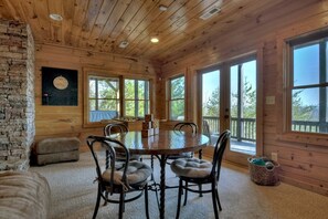 A wooden cabin interior with a round dining table, cushioned chairs, a stone fireplace, and large windows.