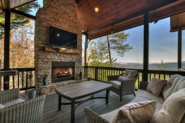 A stone fireplace with a flat-screen TV above it, surrounded by wicker furniture and a wooden deck overlooking a mountainous landscape.