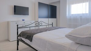 Quarto moderno com cama, televisão na parede, guarda-roupa e janela com cortinas leves.