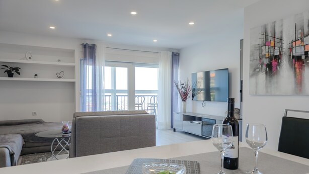 Uma sala de estar moderna com um sofá, uma mesa de jantar com uma tigela de vidro e copos de vinho, uma televisão e uma varanda com guarda-corpo.