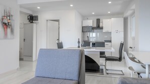 Uma sala de estar moderna e bem iluminada, com um sofá cinzento, área de jantar com uma mesa e cadeiras brancas, e cozinha com armários brancos e eletrodomésticos de aço inoxidável.