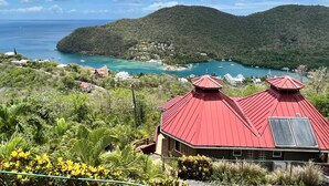 Blick auf die Küste mit einem Haus mit rotem Dach und Solaranlage, umgeben von üppiger Vegetation und blauem Himmel.
