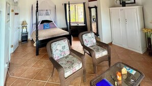 Ein Zimmer mit hoher Holzbalkendecke, einem Himmelbett, zwei Korbsesseln, einem Glastisch mit einer blauen Mappe und einem weißen Schrank.