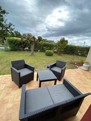 An outdoor patio area with wicker furniture, a small table, and a view of a garden with a tree and bushes.