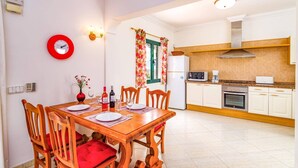 A dining area with a wooden table and chairs, a red clock, and a kitchen with white cabinets and appliances. - Villa Atlantico Mar by Villa Plus (Tías)