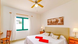 A bedroom with a bed, a nightstand, a chair, a ceiling fan, and a window with curtains. - Villa Atlantico Mar by Villa Plus (Tías)