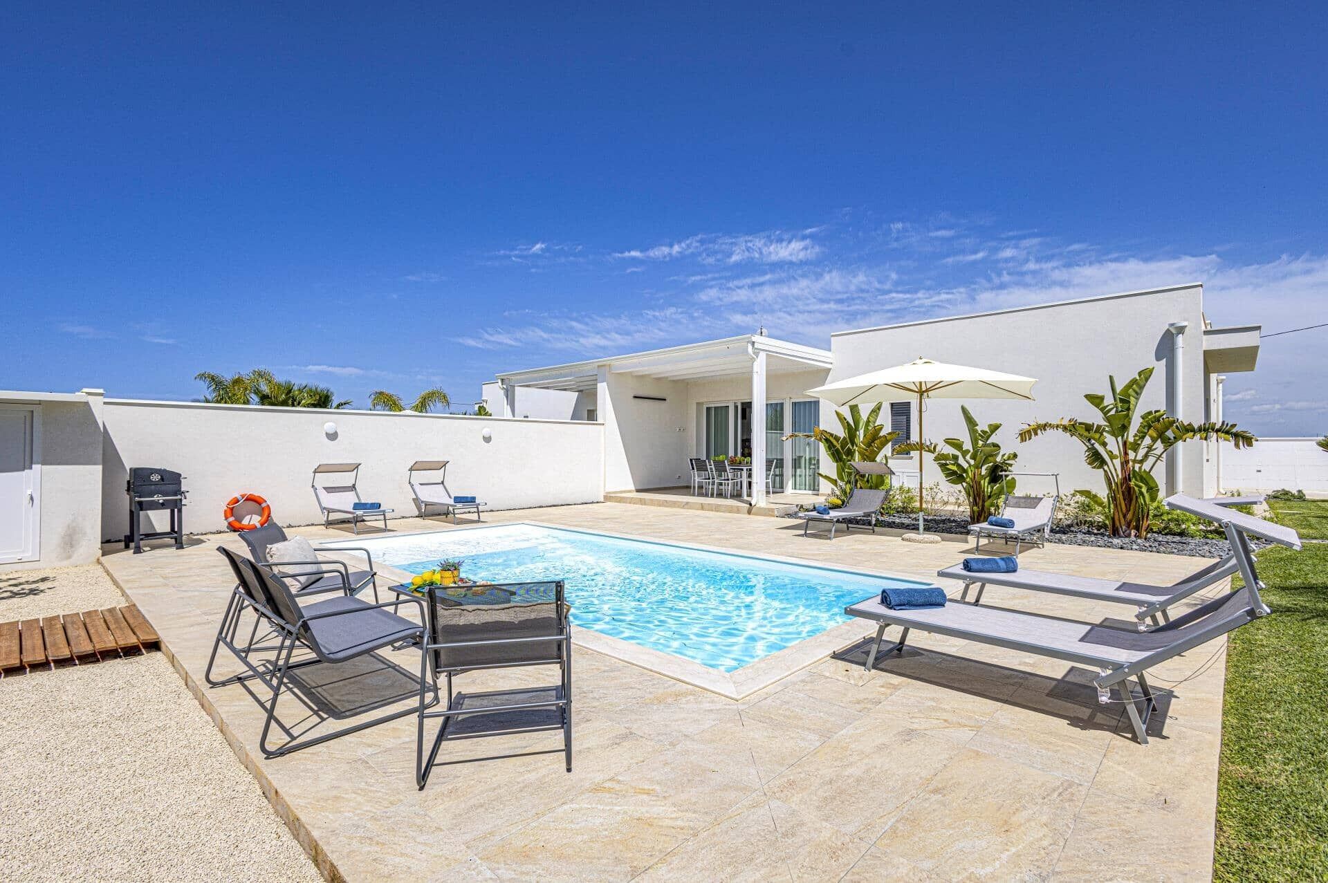 A modern outdoor pool area with lounge chairs, a barbecue, and a covered patio.