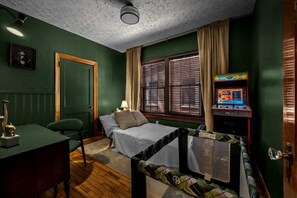 A room with a bed, a desk, a chair, a window with blinds, and a small arcade machine. - San Marco 2BR • King • Porch & Pool Table (Jacksonville)