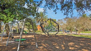 Ein Spielplatz mit Schaukeln und einer Kletterkonstruktion, umgeben von Bäumen und einem Gebäude im Hintergrund.