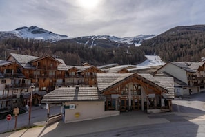 A cluster of wooden buildings with balconies, likely a hotel or resort, set against a backdrop of snow-covered mountains and a forested valley.