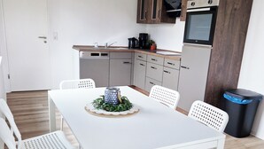 A compact kitchen with wooden cabinets, a built-in oven, and a coffee maker. There is a dining table with white chairs and a decorative centerpiece. - Weser Pension - Zweibettzimmer mit Eigenem Bad Nr. 1 (Brake (Unterweser))