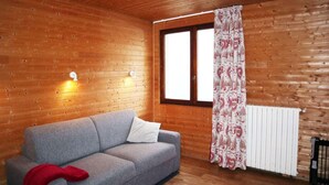 A wooden cabin room with a grey sofa, two wall-mounted lights, a window with a patterned curtain, and a radiator. - 100m from ski slopes, 1st floor, view valley, tv, ski locker, 20m², Les Orres (Les Orres)