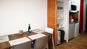 Uma sala de jantar com uma mesa de madeira, cadeiras e uma garrafa de vinho. É visível uma cozinha com micro-ondas e frigorífico ao fundo.