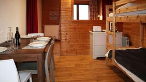 Une chambre confortable, aménagée dans le style d’un chalet, avec une table à manger en bois pour quatre personnes, un lit superposé doté d’un petit bureau et d’un radiateur, et une fenêtre avec des rideaux.