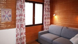 Une pièce chaleureuse avec un canapé gris, des rideaux à motifs, un mur en bois et un panneau orné d’un symbole représentant des skis.
