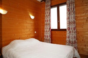A wooden cabin room with a bed, two wall-mounted lights, and a window with patterned curtains. - 100m from resort center, 3rd floor, balcony, tv, ski locker, 80m², Les Orres (Les Orres)