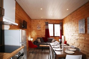 A cozy cabin interior with a wooden interior, a dining table set for two, a sofa, and a kitchen area with a refrigerator.