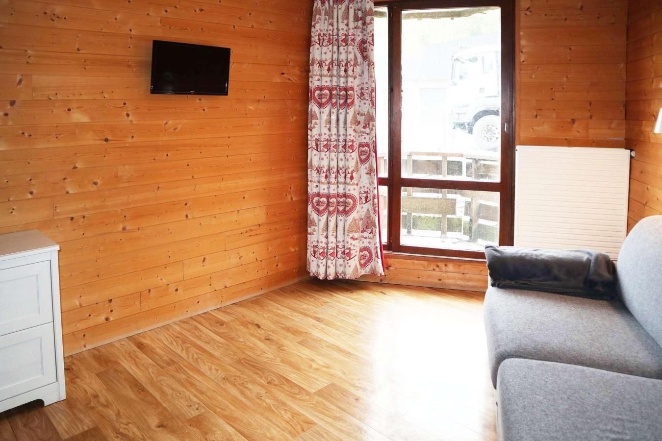 Ein Zimmer mit Holzwänden, einem weißen Schrank, einem grauen Sofa und einem Fenster mit Vorhang.
