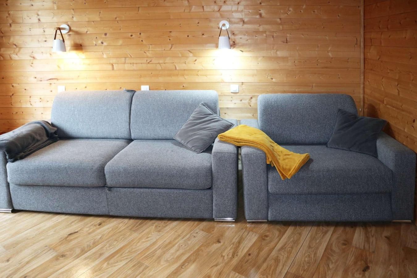 A grey sofa with a throw blanket and pillows, a wooden floor, and two wall-mounted lamps.