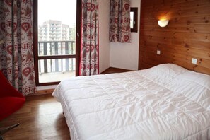 A hotel room with a bed, a red chair, a window with patterned curtains, and a wooden wall with a light fixture.