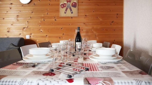 Una mesa de comedor dispuesta con platos blancos, vasos, cubiertos, una botella de vino y un mantel a cuadros rojo y blanco.