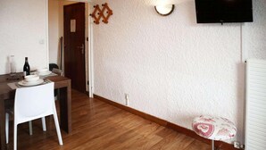 Une pièce avec un plancher en bois, une table à manger avec des chaises, un téléviseur fixé au mur et un radiateur.