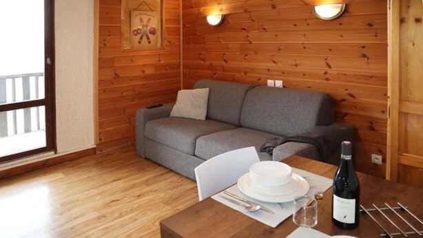 Une pièce chaleureuse avec un plancher en bois, un canapé gris et une table à manger dressée avec des assiettes, des couverts et une bouteille de vin.