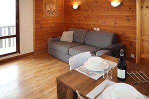 A cozy room with a wooden floor, a gray sofa, a dining table set with plates, cutlery, and a bottle of wine.