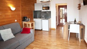Un espace de vie compact comprenant un canapé gris, une petite table en bois, un coin cuisine et un espace repas avec une chaise blanche.
