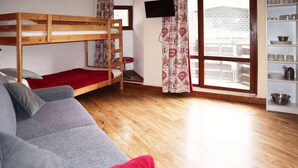 A room with a bunk bed, grey sofa, wooden floor, window with curtains, and a shelf with kitchenware.