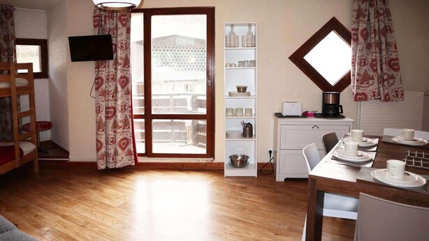A cozy room with a wooden floor, a bunk bed, a dining table set with white plates and cups, a white cabinet with glassware, and a window with patterned curtains.
