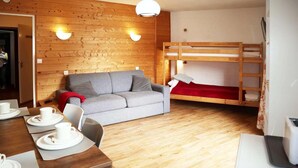 A cozy room with a wooden floor, a grey sofa, a bunk bed, a dining table with plates and cups, and a lamp.