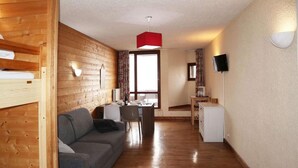 Una habitación acogedora con una pared de madera, un sofá gris, una mesa de comedor con sillas, un refrigerador pequeño y un televisor en la pared.