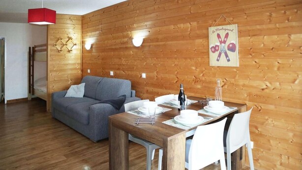 Una habitación acogedora con una pared de madera, un sofá gris, una mesa de comedor de madera con platos blancos y una botella, y una lámpara colgante roja.