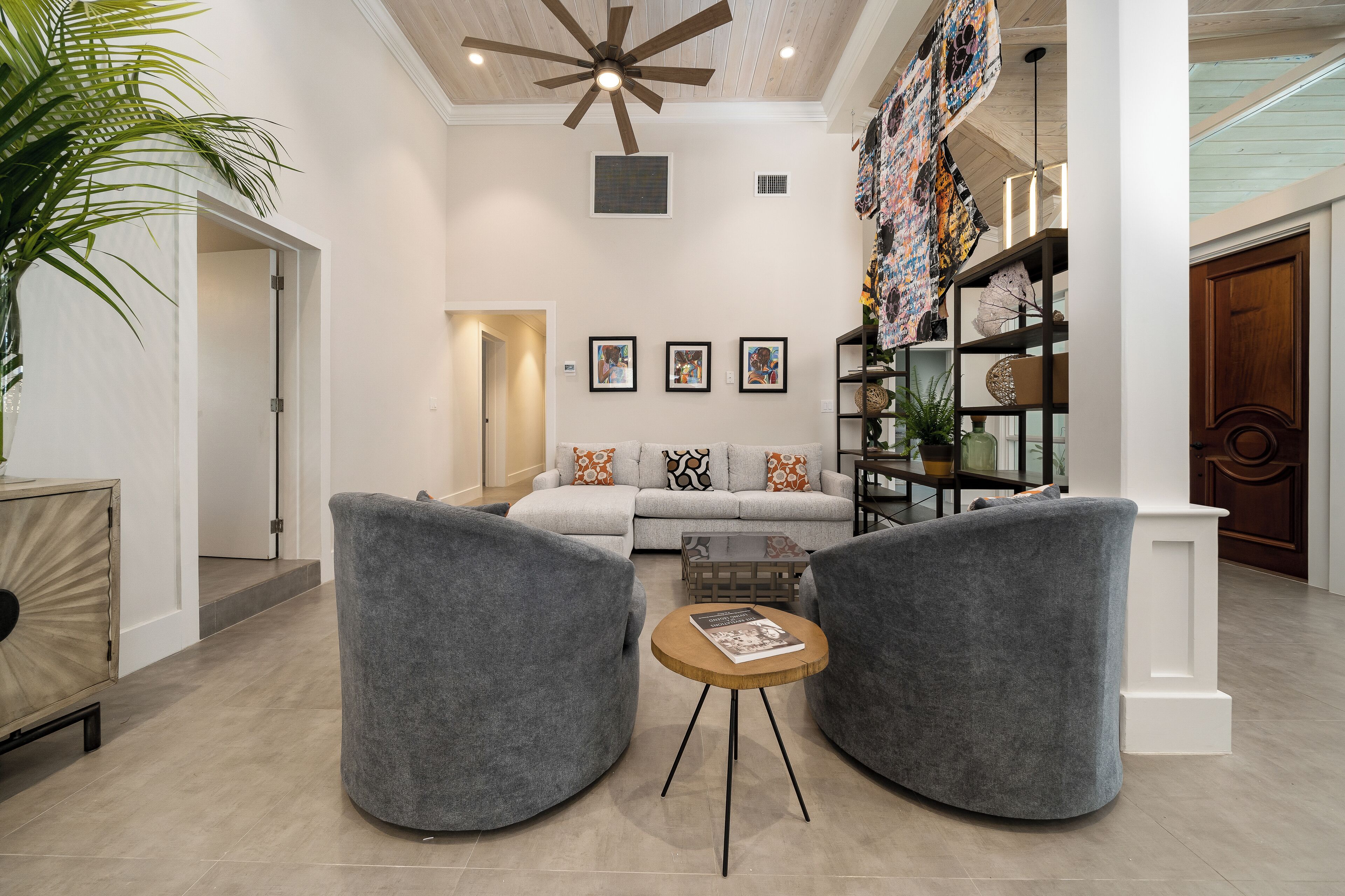 A modern living room with a grey sofa, two grey armchairs, a wooden coffee table, and a ceiling fan. There are framed pictures on the wall, a bookshelf with decorative items, and a large plant.