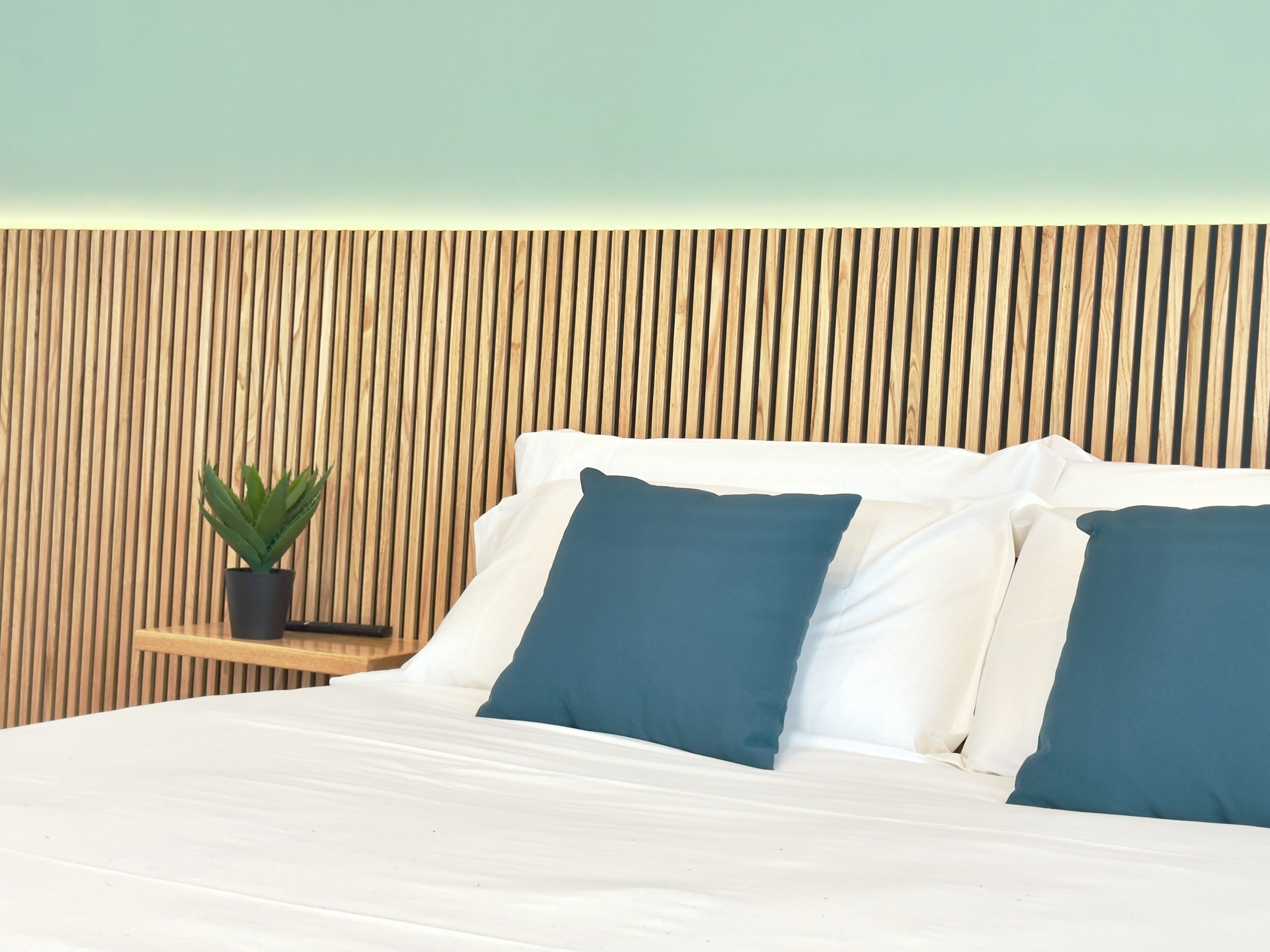 A bed with white and blue pillows, a small wooden nightstand with a plant, and a wooden slatted headboard.