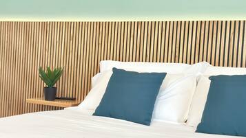 A bed with white and blue pillows, a small wooden nightstand with a plant, and a wooden slatted headboard.