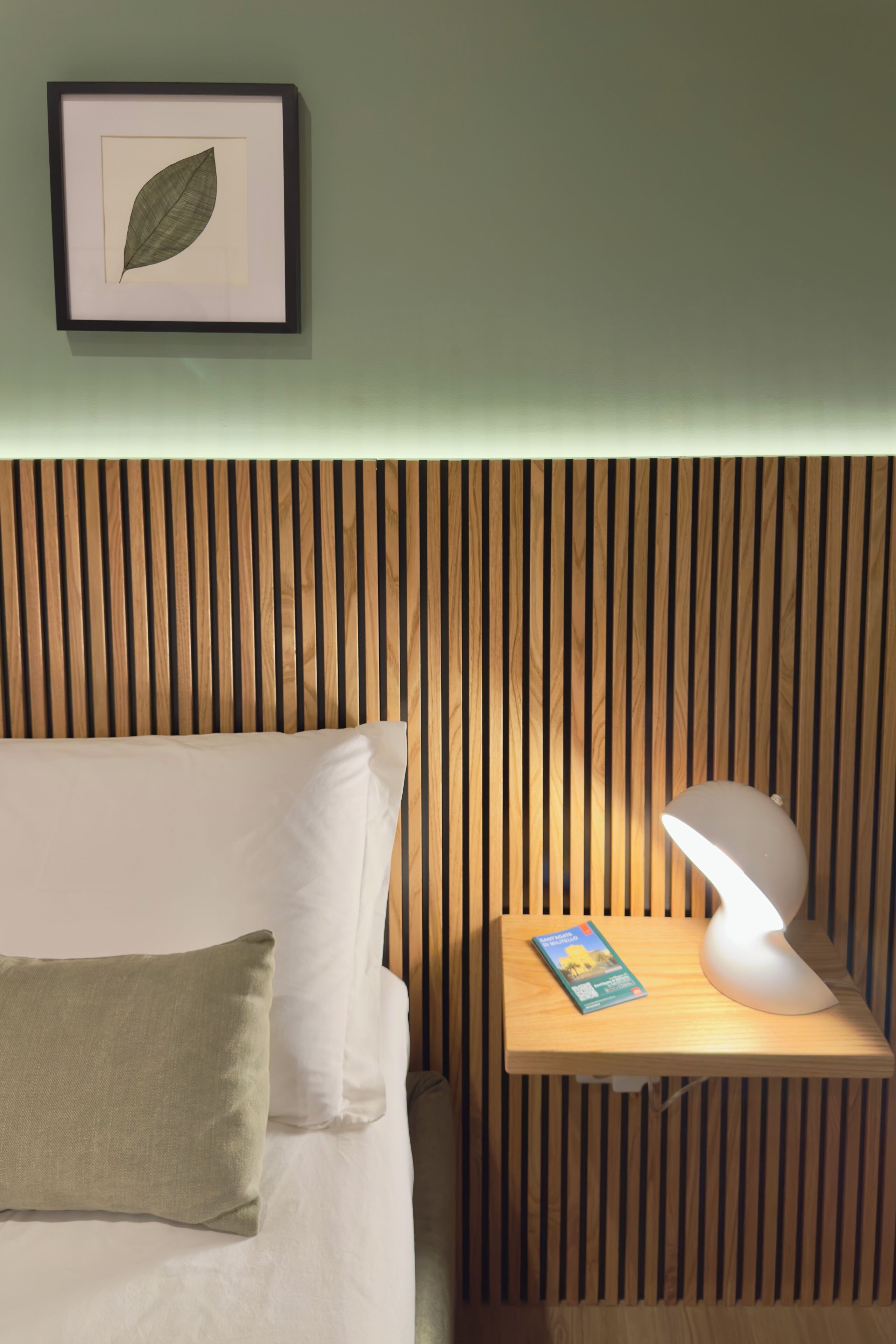 A neatly made bed with pillows, a bedside table with a lamp and a book, and a framed leaf on the wall.