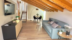 Un salon moderne avec un canapé, une table basse et un téléviseur fixé au mur. Un escalier en bois mène à un niveau supérieur.