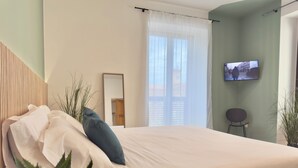 Une chambre à coucher moderne avec un grand lit, un téléviseur fixé au mur et une fenêtre ornée de rideaux légers.