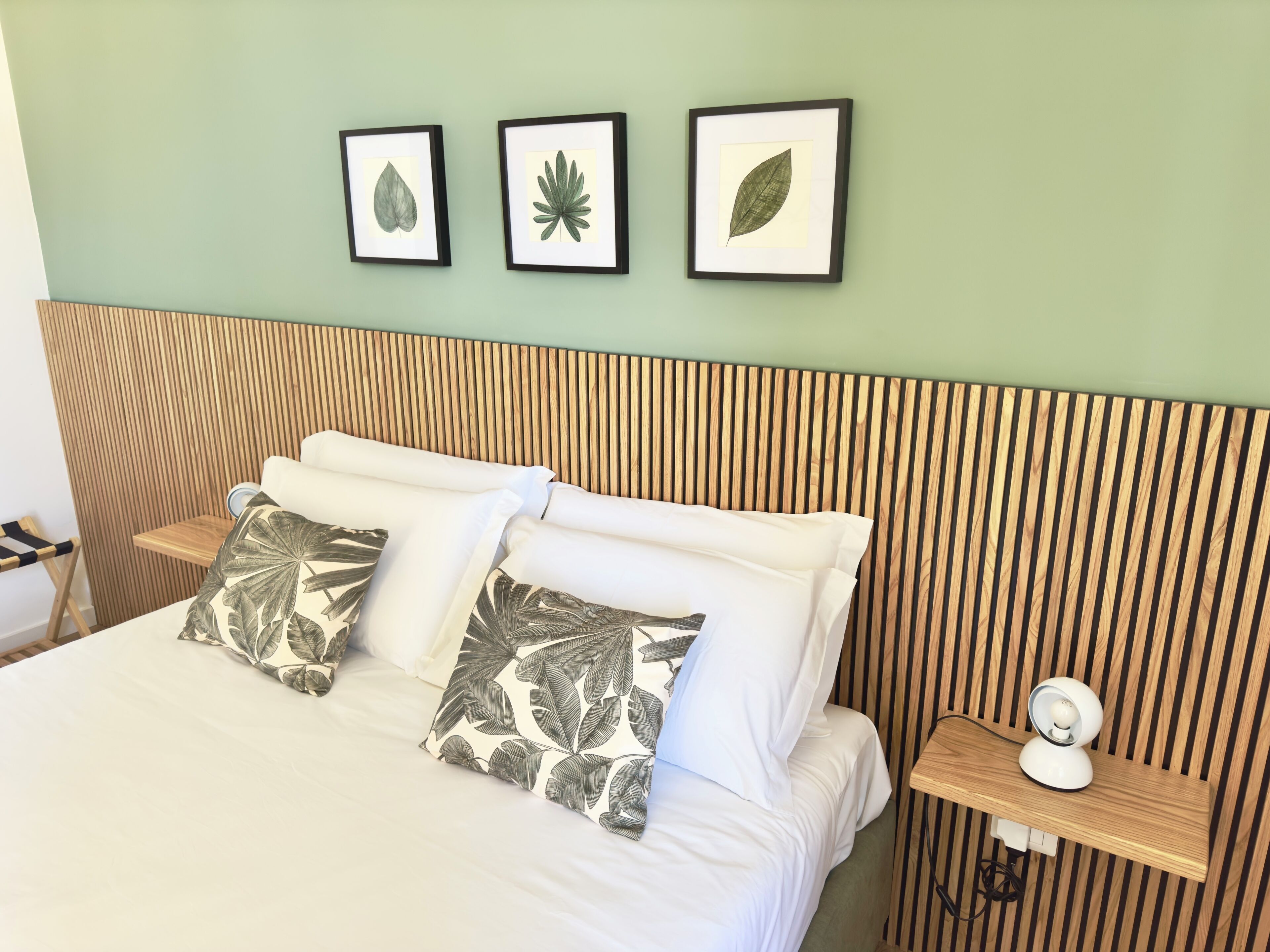 A neatly made bed with white linens and leaf-patterned pillows, a wooden slat headboard, and three framed botanical prints on the wall.